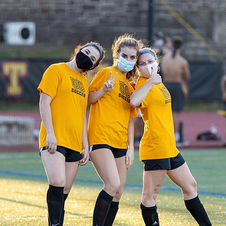 Photo of three Ursinus girl soccer players with masks on standing on the field.