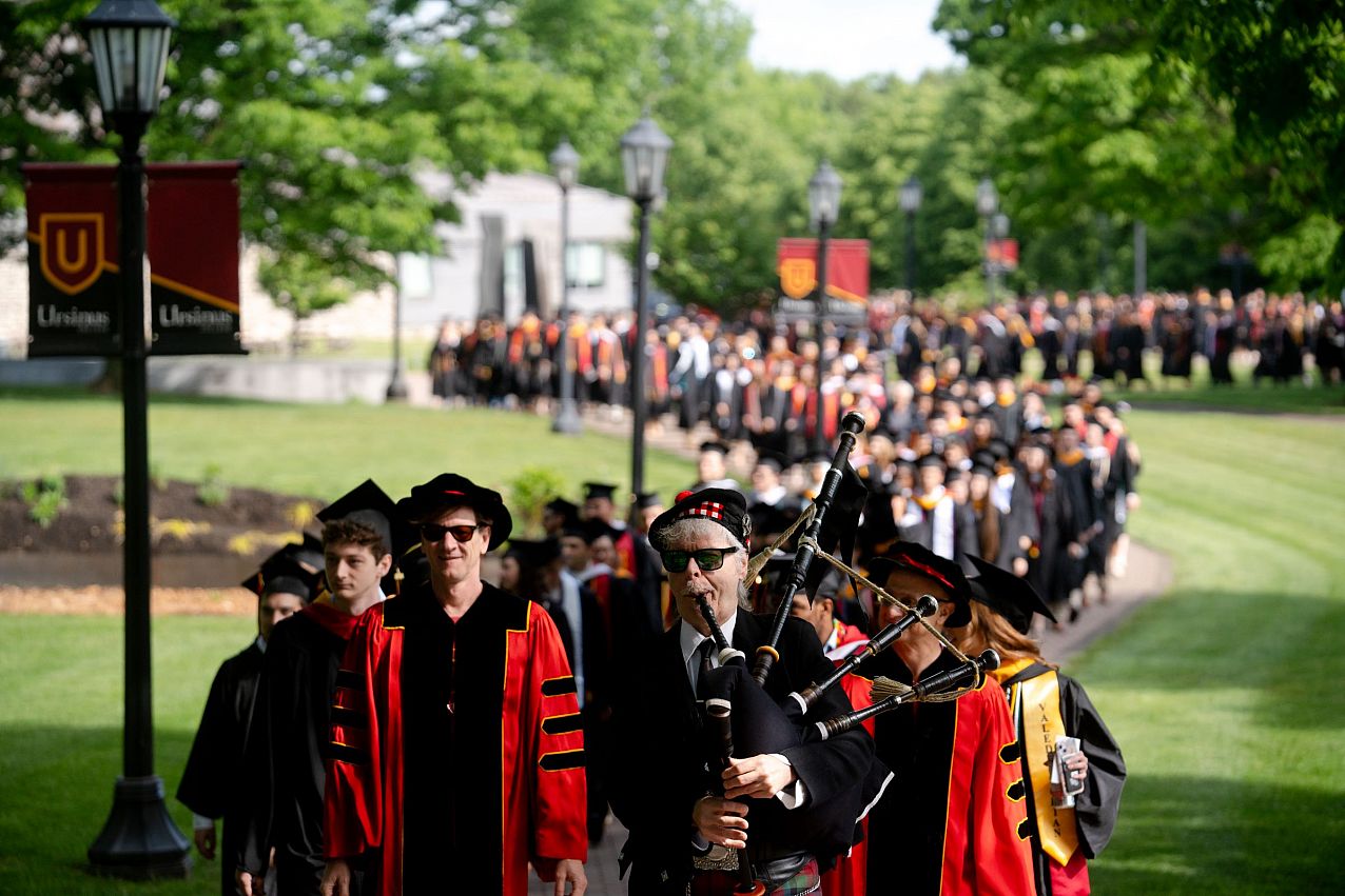 Line of facility and students walking to their commencement ceremony dressed in proper cap and gown attire in an orderly fashion.