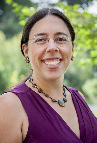 A photo of Dr Leslie New smiling wearing a purple sleeveless top and a green jasper necklace against an out of focus background of green ...