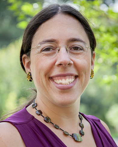 A photo of Dr Leslie New smiling wearing a purple sleeveless top and a green jasper necklace against an out of focus background of green ...