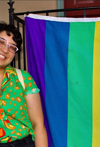 Photograph of a non-binary person standing next to a pride flag.