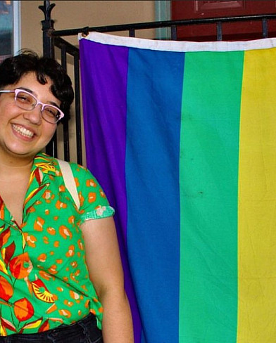 Photograph of a non-binary person standing next to a pride flag.